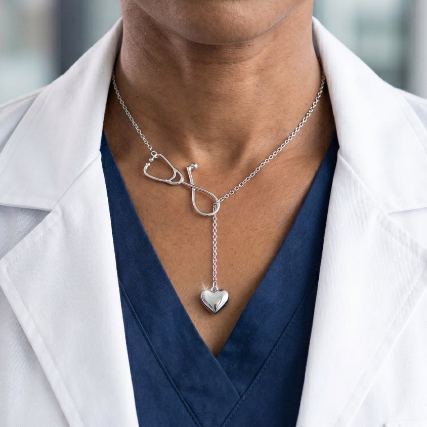 Person wearing a silver necklace with a heart and stethoscope pendant, dressed in a white coat and blue top.